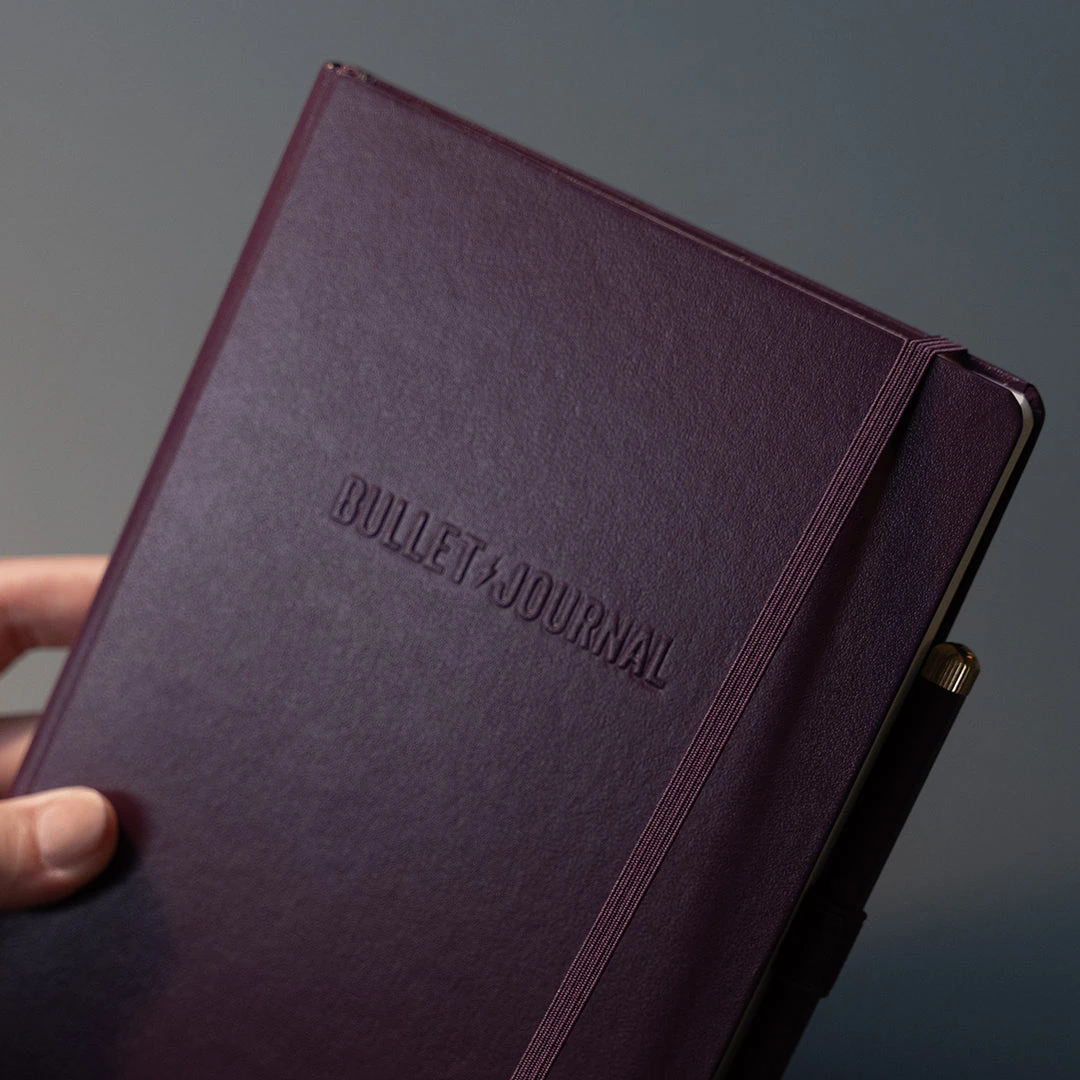 The Purple26 Bullet Journal hardcover held in the hand, showing its embossed cover and elastic closure